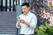 © Liubomir - Happy and confident Asian businessman typing a message on the phone outside the office