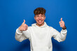 © Andres - Attractive cheerful young man with curly hair wearing a white sweatshirt standing isolated over blue background, showing thumbs up while smiling and looking at the camera.