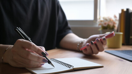 Wall Mural - Close up stylish man hand writing on notebook and using mobile phone at home office desk.