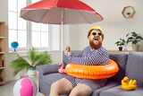 Fototapeta  - Summer party at home. Funny chubby man having fun sitting on sofa in living room with inflatable swimming circle. Humorous man has absurd vacation under beach umbrella on improvised home beach.
