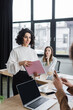 © LIGHTFIELD STUDIOS - Smiling muslim businesswoman holding paper folder near devices and multiethnic colleagues in office.