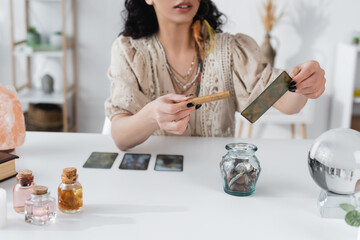 Wall Mural - Cropped view of fortune teller holding sage stick and tarot card at home.