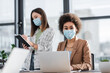 © LIGHTFIELD STUDIOS - African american businesswoman in medical mask looking at camera near laptop and colleague in office.