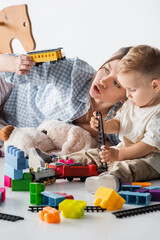 young mother holding toy train near playing son on white.