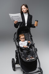 businesswoman with coffee to go reading newspaper near son in baby carriage on grey.