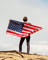 A woman with an American flag stands on the sand, skies on the background. Woman is holding with both hands waving American USA flag. Fourth of July Independence Day.