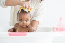 Cute African Newborn Baby Bathing In Bathtub With Soap Bubbles On Head And Body. Mother Use Sponge With Shampoo Or Shower Gel To Wash Little Daughter In Water. Newborn Baby Cleanliness Care Concept