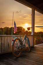 Vertical Shot Of A Bike Outside Of A Wooden House By The United State Flag Against The Sunset Sky