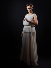 Wall Mural - An ancient heroine, a young woman in the image of an ancient Greek goddess or muse. A noble heroine in a white tunic and a laurel wreath, a full-length photo on a black background