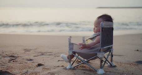 Wall Mural - happy cute adorable Asian baby infant sitting relaxing on little chair with waves on background at seaside tropical sandy beach in sunrise during holiday vacation summertime Thailand	
