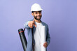 Young architect Moroccan man with helmet and holding blueprints over isolated background pointing front with happy expression