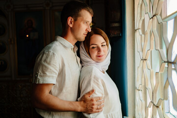 man and woman in headscarf and light-colored robes by window in church. 