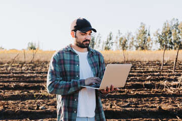 Young latin farmer man teleworking on his laptop in the middle of his farmland . Agricultural sustainability