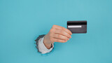 Fototapeta  - A woman's hand sticking out of a hole in a paper blue background holds a credit card. 