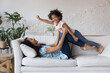 © fizkes - Happy African mom lifting joyful daughter girl in air, lying on soft couch, laughing, doing acroyoga support with kid in arms. Excited mother and flying superhero child playing active game