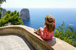 Beautiful young woman with hat sitting on wall looking at stunning panoramic view of Capri Island with Faraglioni sea stack and blue crystalline water on the background, Capri, Italy
