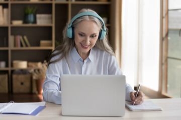 Wall Mural - Happy mature training course student woman in wireless earphones watching webinar on laptop computer, writing notes, studying on Internet, making video call, chatting online, smiling