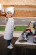 © Aleksandr - Adorable little girl in chef's coat and cap cooks at the children's toy kitchen. Playing on little kids city. Girl thumbs up to the camera
