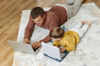 © KIFOR PRODUCTION - Dad and son are lying on the floor with computers. Dad teaches his son programming. Children's computer.