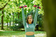© nikjvt - Attractive young brunette girl in olive green sports outfit lifting fitness weights facing forward during the outdoor workout exercise near the public fountain in the urban city forest like park