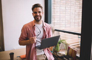 Wall Mural - Half length of happy male freelancer with laptop device and spectacles in hands smiling at camera during work break, joyful Caucaisan programmer with digital netbook enjoying time for networking