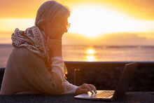 Silhouette Of Senior Active Woman Close To The Beach Using Laptop Wearing Eyeglasses. Elderly Attractive Female In Remote Work Enjoying Orange Sunset At Sea, Horizon Over Water