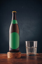 Brown Beer Bottle With Green Blank Label And Empty Glass On Wooden Table, Dark Backdrop. Alcohol Beverage Product Advertising