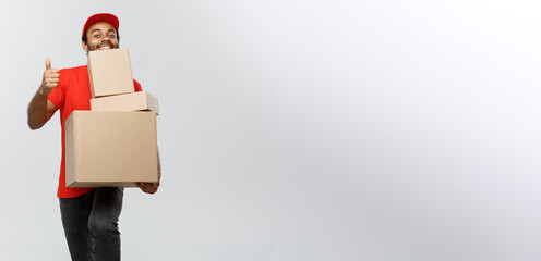 Wall Mural - Delivery Concept - Portrait of Happy African American delivery man holding box packages and showing thumps up. Isolated on Grey studio Background. Copy Space.