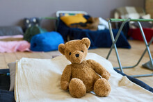 Teddybear Of Brown Color Sitting On Bed Done With White Cotton Blanket Of Refugee Child In Large Room Prepared For Migrants