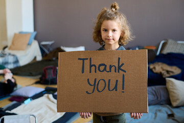 Wall Mural - Cute blond little boy with cardboard poster saying thank you looking at camera while standing against sleeping places in refugee camp