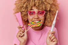 Oral Hygiene And Teeth Care Concept. Surprised Curly Haired Woman With Multicolored Candy In Mouth Wears Heart Shaped Pink Sunglasses Holds Toothpaste And Electric Toothbrush Isolated On Pink Wall