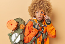 Excited Shocked Woman With Curly Hair Holds Magnifying Glass Over Eye Has Rest During Travel Carries Heavy Rucksack Has Journey Isolated Over Brown Background. People And Recreation Concept.