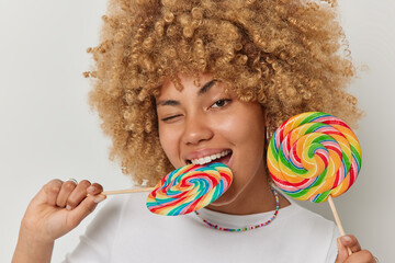 Wall Mural - Headshot of curly haired beautiful woman bites multicolored caramel candy on stick winks eye has glad expression enjoys eating delicious sweet rainbow lollipops poses against white background.