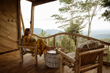 Young  pretty tourist girl sit on terrace of wooden eco house with mountains view and drink coconut. Enjoy vacation on Bali island