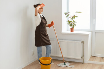 Full length portrait of tired dark haired woman wearing white t shirt and apron posing with mop, being exhausted, keeping hand on her forehead, standing near window.