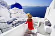 © zigres - Beautiful girl in red dress and hat comes down the stairs to her resort enjoying a spectacular view of the Caldera in Oia village, Santorini, Greece