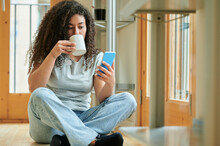 Young Woman Drinking Coffee And Using Mobile Phone Sitting On Floor At Home