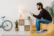 © Westend61 - Happy man with beard using mobile phone sitting on sofa in living room at home