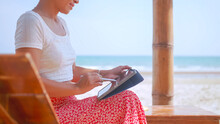 Asian Woman Drawing On A Tablet While Sitting By The Sea. Freelancer Working On The Beach.