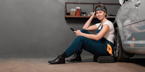 Wall Mural - Banner of women's equality. Smiling caucasian young woman mechanic is sitting leaning on a car in an auto repair shop and using cellphone. Copy space. The concept of women's work in male professions