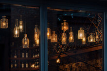 Lanterns Made Of Glass Bottles And Edison Lamp Bulbs. DIY Lamps Made From Recycled Old Bottles Hanging In Cafe Window.