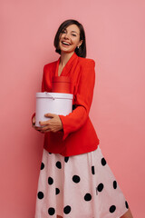 Wall Mural - Cute young caucasian girl smiles, holds gift box in her hands on pink background. Brunette with short haircut wore red jacket and white skirt for occasion. Celebration concept