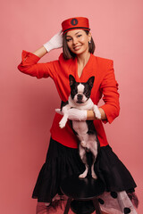 Wall Mural - Beautiful young caucasian woman holding purebred dog, feeling happy on pink background. Brunette girl in red suit looks at camera. Animal owner concept