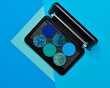 Blue eyeshadow makeup palette. Realistic black case with matte and glitter pigments. Shadows set in colors of ocean wave. Top view flat lay package isolated on blue background.