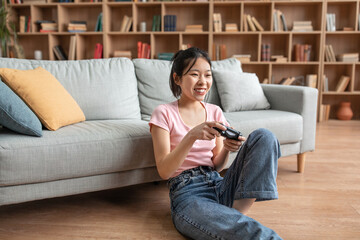 Canvas Print - Happy asian lady holding joystick and playing videogames while resting on sofa at home, free space