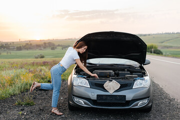 Wall Mural - A young girl stand near a broken car in the middle of the highway during sunset and tries to repair it. Troubleshooting the problem. Car service. Car breakdown on the road.