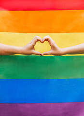 close up of two woman's hands forming a heart with the lgbtq rainbow flag in the background. symbol 
