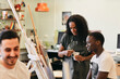 © Maskot - Female tutor looking at painting on easel by male student sitting in art class