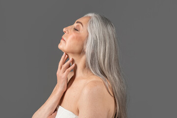 Feminine senior lady with silky skin wearing towel, touching her neck on grey studio background, side view portrait