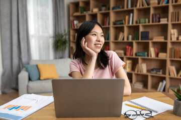 Wall Mural - Portrait of happy dreamy asian lady sitting at desk with laptop at home, looking away, thinking and smiling, free space